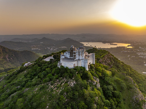 Aerial view of Sajjangarh Monsoon Palace perched on a lush green hilltop overlooking the surrounding landscape and a distant lake at sunset in Udaipur Rajasthan India.