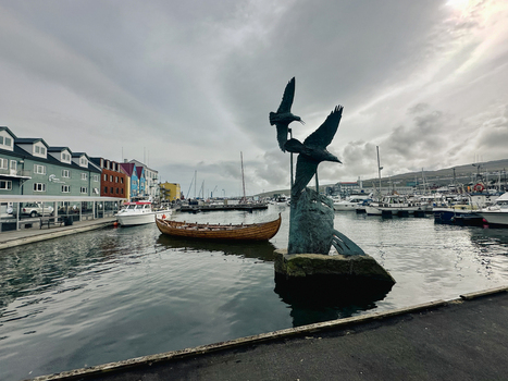 Faroe Islands 28 August 2024 View of the harbor in Torshavn with a bronze bird sculpture a traditional wooden boat and colorful buildings under a cloudy sky.