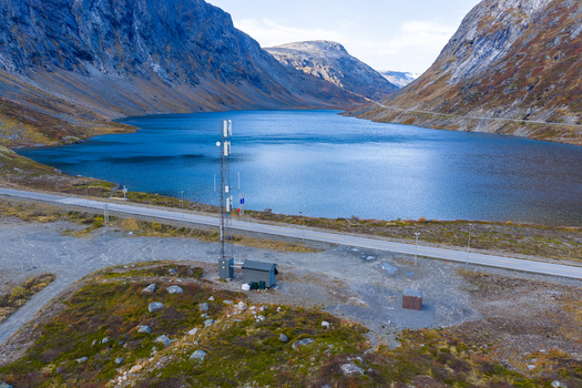 Aerial view of a telecommunications tower situated near a mountain lake and a road surrounded by rugged hills and autumn vegetation in Skjak Innlandet Norway.