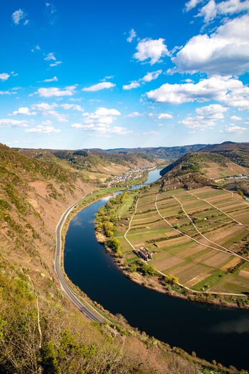 Aerial view of the Moselle River loop winding through terraced vineyards and lush green hills under a bright blue sky with scattered clouds in Bremm Rhineland Palatinate Germany. Aerial view of the Moselle River loop winding through terraced vineyards and lush green hills under a bright blue sky with scattered clouds in Bremm Rhineland Palatinate Germany.