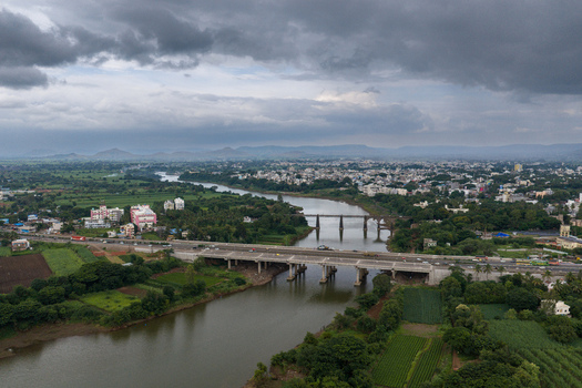 Aerial view of the Krishna River flowing through the town of Karad with bridges lush green agricultural fields and urban buildings under a cloudy sky in Karad Maharashtra India.