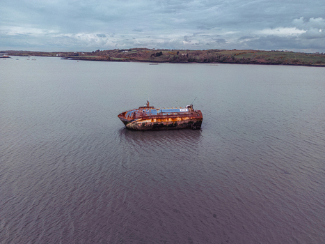 Aerial view of a rusty shipwreck floating in calm water near a distant green shoreline under an overcast sky. Aerial view of a rusty shipwreck floating in calm water near a distant green shoreline under an overcast sky.