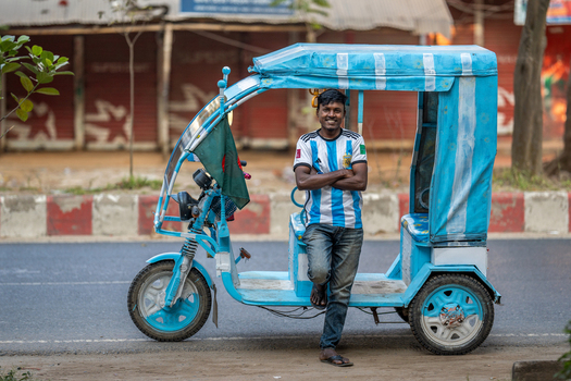 Bogura Bangladesh 02 December 2022 View of a man wearing an Argentina soccer jersey standing next to his blue electric rickshaw on a street in Bogura.