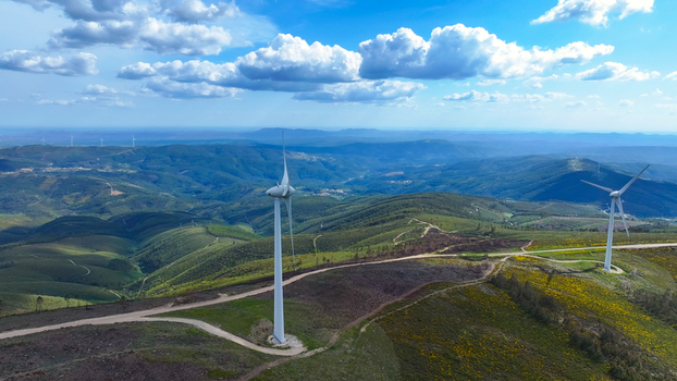 Aerial view of wind turbines situated on rolling green hills with dirt access roads under a bright blue sky with scattered clouds in Lisbon Lisbon Portugal. Aerial view of wind turbines situated on rolling green hills with dirt access roads under a bright blue sky with scattered clouds in Lisbon Lisbon Portugal.