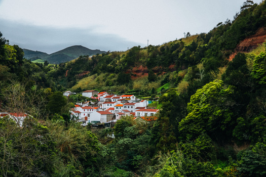 View of white houses with red roofs nestled in a verdant valley contrasting against the rugged terrain under an overcast sky Furnas Azores Portugal.