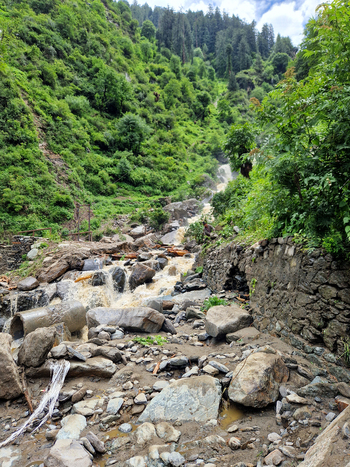 View of the Malana stream flowing through a rocky mountain landscape with lush green hills and debris in Malana Himachal Pradesh India.