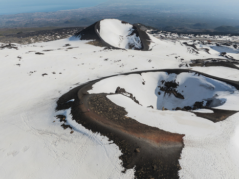Aerial view of the snow covered volcanic craters on the south side of Mount Etna with dark volcanic ash and a distant view of the coastline in Nicolosi Sicilia Italy. Aerial view of the snow covered volcanic craters on the south side of Mount Etna with dark volcanic ash and a distant view of the coastline in Nicolosi Sicilia Italy.