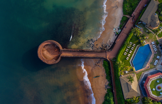 Aerial view of the historic Fort Aguada stone bastion extending into the turquoise sea with a sandy beach and a luxury resort pool Candolim Goa India.