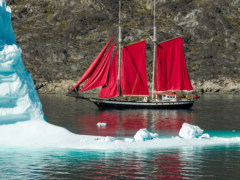 Aerial view of a sailboat with striking red sails navigating near a large iceberg in the icy waters of Greenland. Aerial view of a sailboat with striking red sails navigating near a large iceberg in the icy waters of Greenland.