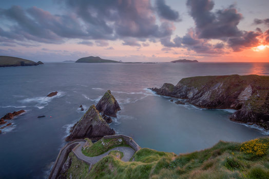 View of the winding stone path of Dunquin pier leading to the Atlantic Ocean with rugged cliffs and islands under a sunset sky Dun Chaoin County Kerry Ireland. View of the winding stone path of Dunquin pier leading to the Atlantic Ocean with rugged cliffs and islands under a sunset sky Dun Chaoin County Kerry Ireland.