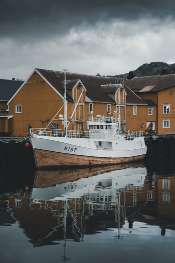 Nusfjord Norway 07 September 2022 View of a fishing boat moored in the harbor of Nusfjord with yellow wooden buildings reflecting in the calm water under a cloudy sky. Nusfjord Norway 07 September 2022 View of a fishing boat moored in the harbor of Nusfjord with yellow wooden buildings reflecting in the calm water under a cloudy sky.