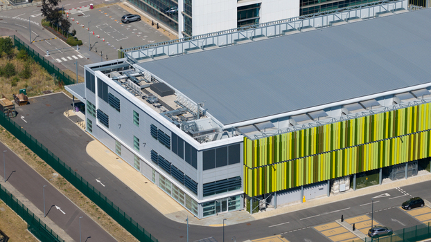 Aerial view of the Kao Data facility with green and yellow facade panels a flat grey roof and surrounding parking areas in Harlow England United Kingdom. Aerial view of the Kao Data facility with green and yellow facade panels a flat grey roof and surrounding parking areas in Harlow England United Kingdom.