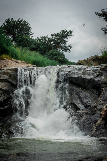 View of a waterfall cascading over dark rocks into a pool surrounded by lush green vegetation and trees under a cloudy sky in Jos North Plateau Nigeria. View of a waterfall cascading over dark rocks into a pool surrounded by lush green vegetation and trees under a cloudy sky in Jos North Plateau Nigeria.