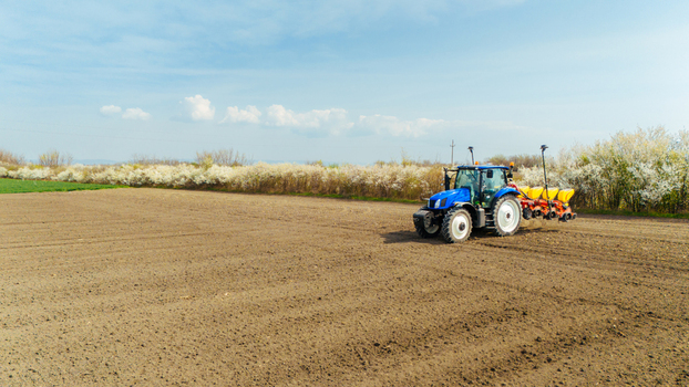 Aerial view of a blue tractor tilling the soil on a vast field contrasted by a row of trees with white blossoms Sremska Mitrovica Vojvodina Serbia. Aerial view of a blue tractor tilling the soil on a vast field contrasted by a row of trees with white blossoms Sremska Mitrovica Vojvodina Serbia.