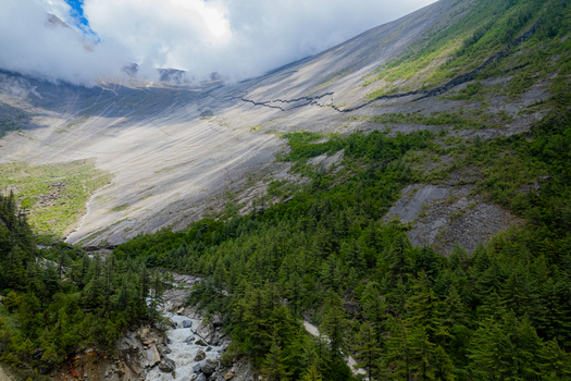 Aerial view of a steep rocky mountain slope with a flowing river and dense pine forests in the valley under a cloudy sky in Nepal.