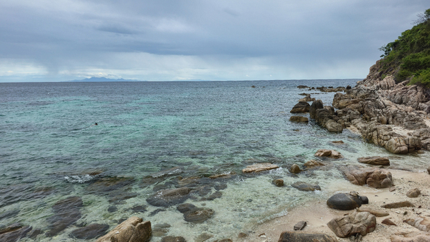 View of the rocky coastline of Ko Tao with clear turquoise water dark storm clouds over the horizon and lush green vegetation on the cliffs in Ko Tao Surat Thani Thailand.