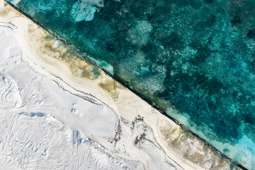 Aerial view of airport project with tire tracks and a dark sea wall barrier bordering turquoise coral reef waters in Makunudhoo Haa Dhaalu Atoll Maldives.