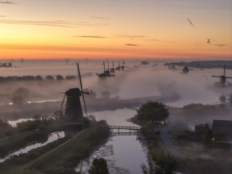 Aerial view of UNESCO World Heritage Kinderdijk windmills shrouded in thick morning mist and fog under a vibrant orange sunrise sky Kinderdijk Zuid Holland Netherlands.