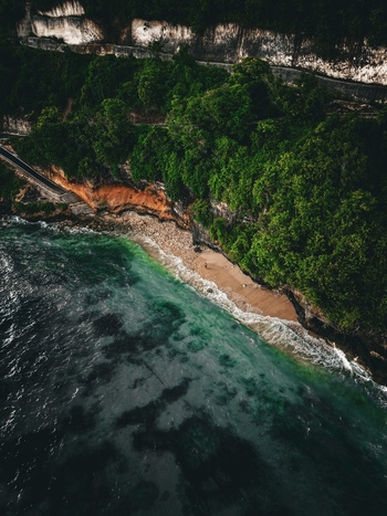 Aerial view of Pantai Nunggalan featuring a secluded beach turquoise waves rugged limestone cliffs with lush vegetation and a coastal road in Kecamatan Kuta Selatan Bali Indonesia.