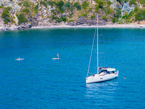 Aerial view of a white sailboat in clear turquoise waters with people on paddleboards nearby and a rocky coastline in the background in Capoliveri Tuscany Italy.