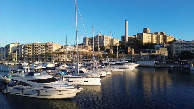 Aerial view of luxury yachts moored in the marina with historic buildings and a tall chimney in the background in Valletta Port Region Malta. Aerial view of luxury yachts moored in the marina with historic buildings and a tall chimney in the background in Valletta Port Region Malta.
