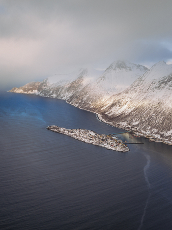 Aerial view of a rugged coastline where snow capped mountains meet the dark swirling sea near a small isolated island Fjordgård Troms Norway. Aerial view of a rugged coastline where snow capped mountains meet the dark swirling sea near a small isolated island Fjordgård Troms Norway.