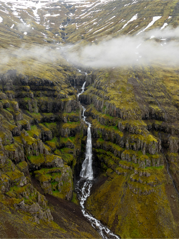 Aerial view of a cascading waterfall flowing down moss covered basalt cliffs with patches of snow and low hanging clouds in the Eastern Region Iceland. Aerial view of a cascading waterfall flowing down moss covered basalt cliffs with patches of snow and low hanging clouds in the Eastern Region Iceland.