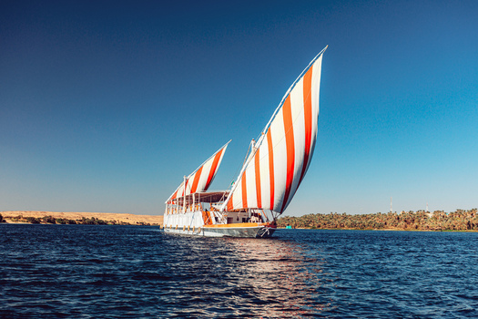 View of a traditional dahabiya sailboat with red and white striped sails cruising on the Nile River under a clear blue sky in Aswan Aswan Governorate Egypt. View of a traditional dahabiya sailboat with red and white striped sails cruising on the Nile River under a clear blue sky in Aswan Aswan Governorate Egypt.