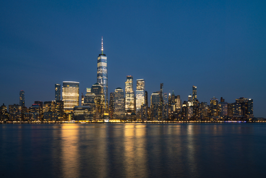 View of the One World Trade Center and the illuminated Manhattan skyline reflecting on the Hudson River at twilight in New York New York United States.