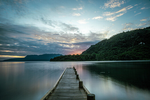 View of a wooden jetty extending into the calm waters of Lake Tarawera at sunset with lush green hills in the background in Rotorua Bay of Plenty Region New Zealand.