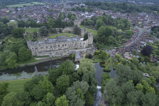 Aerial view of Warwick Castle surrounded by lush green trees and the River Avon with the historic town in the background in Warwick England United Kingdom. Aerial view of Warwick Castle surrounded by lush green trees and the River Avon with the historic town in the background in Warwick England United Kingdom.