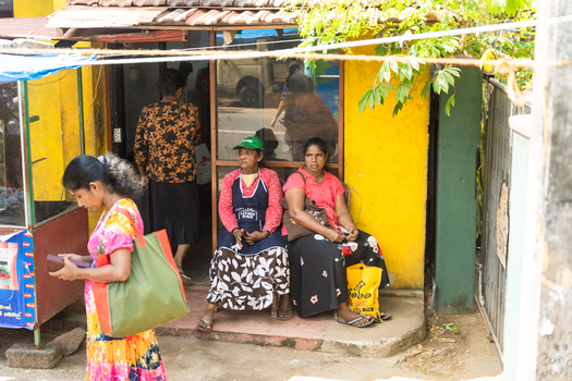 Sri Lanka 31 January 2026 View of local women sitting outside a small yellow building with a woman walking in the foreground.