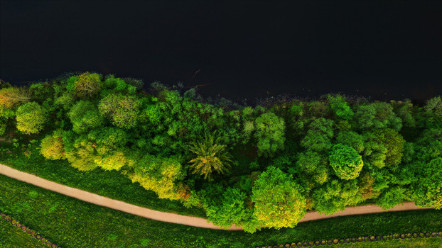 Aerial view of a lush green riverbank with dense trees a winding walking path and dark water in Latvia. Aerial view of a lush green riverbank with dense trees a winding walking path and dark water in Latvia.