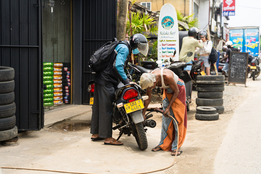 Sri Lanka 18 February 2026 View of an elderly man in a sarong inflating a motorcycle tire for a delivery driver at a roadside shop with stacks of tires and a surf school sign.