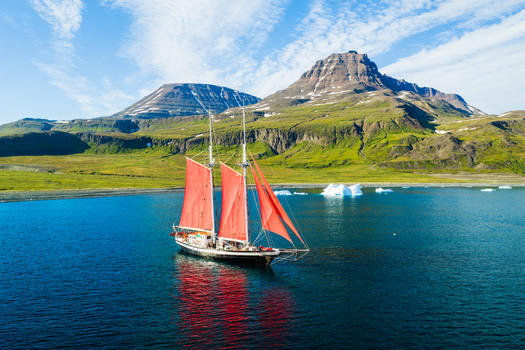 Aerial view of a sailboat with vibrant red sails navigating through calm blue waters surrounded by dramatic green mountains and floating icebergs in Greenland. Aerial view of a sailboat with vibrant red sails navigating through calm blue waters surrounded by dramatic green mountains and floating icebergs in Greenland.