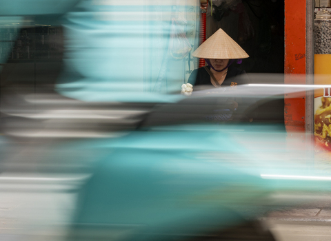 View of a person wearing a traditional conical hat sitting on a street with a blurred scooter passing by in Hanoi Ha Noi Vietnam.