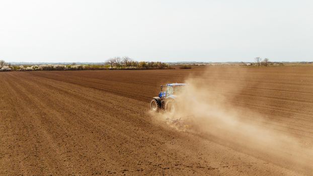 Aerial view of a lone blue tractor churning up the rich brown soil kicking up dust under a muted sky in the fields Sremska Mitrovica Vojvodina Serbia. Aerial view of a lone blue tractor churning up the rich brown soil kicking up dust under a muted sky in the fields Sremska Mitrovica Vojvodina Serbia.