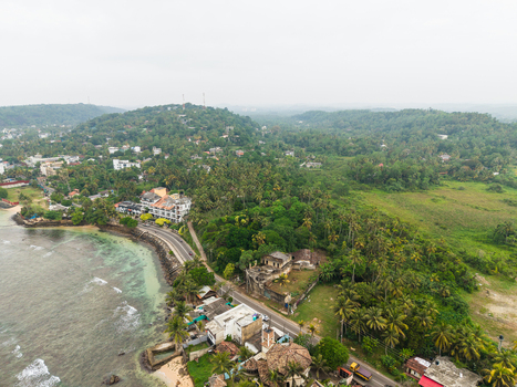 Aerial view of the coastal road winding along the turquoise ocean and dense tropical forest with palm trees and scattered buildings in Unawatuna Southern Province Sri Lanka.