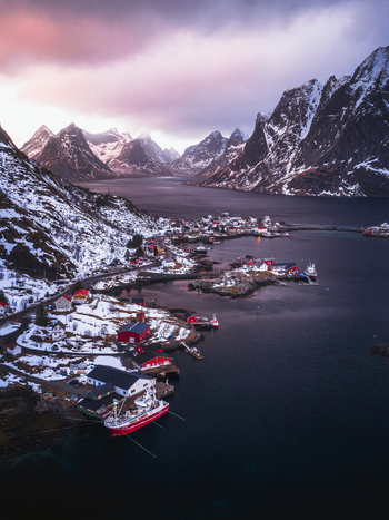 Aerial view of vibrant red cabins nestled against snow dusted mountains reflecting in the tranquil waters of the fjord Reinebringen Nordland Norway.