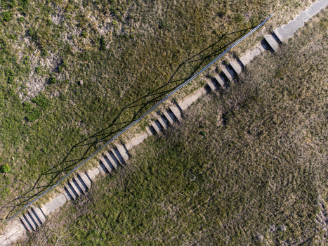 Aerial view of a concrete path bisecting the landscape casting stark shadows beside a dark fence line contrasting with the muted greens and browns Almere Flevoland Netherlands.