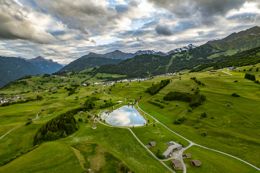 Aerial view of the village of Prutz surrounded by lush green meadows a small lake and rolling hills under a cloudy sky in Prutz Tyrol Austria. Aerial view of the village of Prutz surrounded by lush green meadows a small lake and rolling hills under a cloudy sky in Prutz Tyrol Austria.
