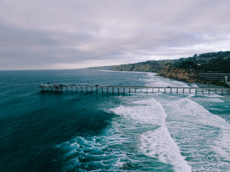 Aerial view of Scripps Pier stretching into the Pacific Ocean with crashing waves and coastal cliffs under a cloudy sky in San Diego California United States. Aerial view of Scripps Pier stretching into the Pacific Ocean with crashing waves and coastal cliffs under a cloudy sky in San Diego California United States.