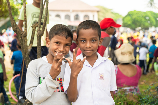 Sri Lanka 12 February 2026 View of two smiling schoolboys making hand gestures during an outdoor event with a crowd and greenery in the background.