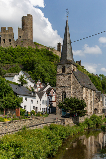View of the historic stone church and half timbered houses along the river with medieval castle ruins on a lush green hill in Monreal Rhineland Palatinate Germany. View of the historic stone church and half timbered houses along the river with medieval castle ruins on a lush green hill in Monreal Rhineland Palatinate Germany.
