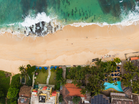 Aerial view of the tropical coastline featuring turquoise ocean waves crashing on golden sand and a resort with palm trees in Unawatuna Southern Province Sri Lanka.