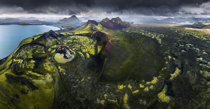 Aerial view of a volcanic crater surrounded by green and yellow moss covered hills dark lava fields and a calm lake under a stormy sky in Iceland.