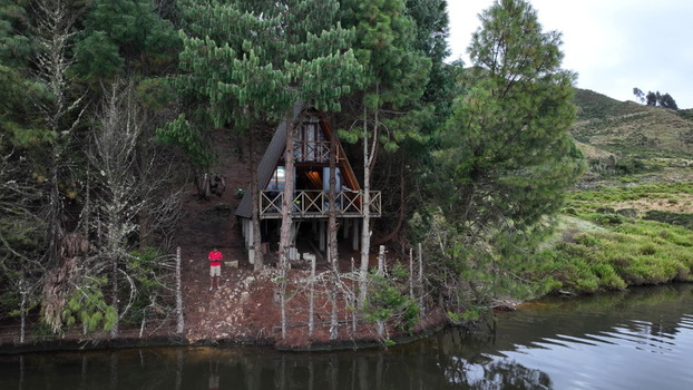 Aerial view of an A frame cabin nestled among pine trees by Corani Lake with a person in a red shirt standing nearby in Chapare Cochabamba Department Bolivia. Aerial view of an A frame cabin nestled among pine trees by Corani Lake with a person in a red shirt standing nearby in Chapare Cochabamba Department Bolivia.