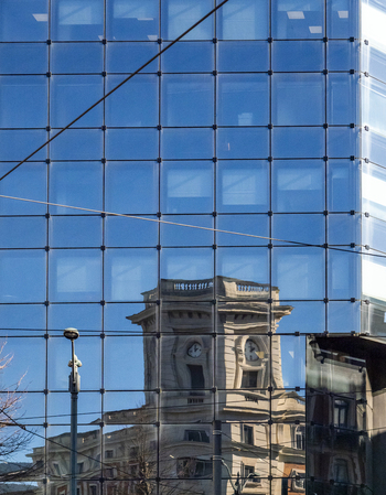 View of the reflection of a historic clock tower building on a modern glass skyscraper facade under a clear blue sky Greater Bilbao Basque Country Spain. View of the reflection of a historic clock tower building on a modern glass skyscraper facade under a clear blue sky Greater Bilbao Basque Country Spain.