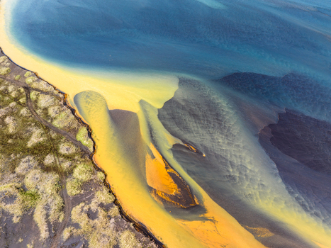 Aerial view of a glacial river delta with vibrant yellow sediment mixing with dark blue glacial water and black volcanic sand against a textured mossy landscape in Iceland. Aerial view of a glacial river delta with vibrant yellow sediment mixing with dark blue glacial water and black volcanic sand against a textured mossy landscape in Iceland.
