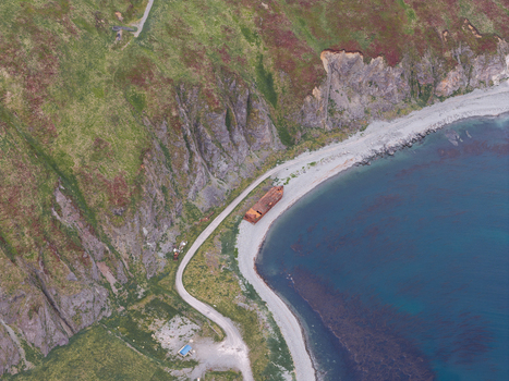 Aerial view of a rusted shipwreck resting on a rocky beach along the coastline with steep green hills and a winding road in Unalaska Alaska United States.